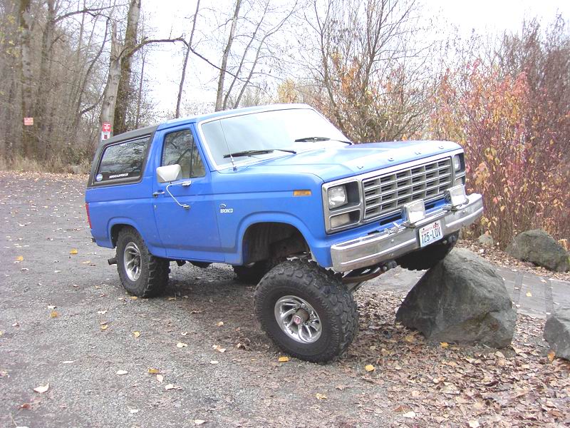 Blue lifted Ford Bronco with front tire on a boulder, front three-quarter view