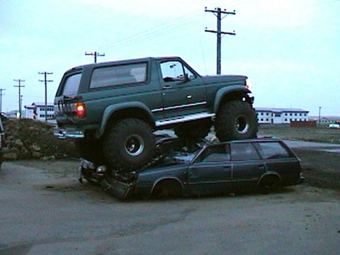 1996 Ford Bronco - A lifted 1996 Ford Bronco is parked on top of a dark station wagon, with a rural landscape in the background.