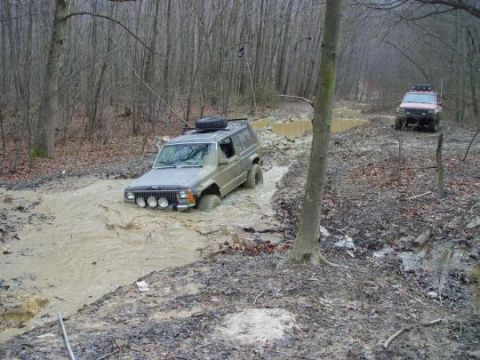 1997 Ford Expedition - Two SUVs navigate a muddy trail through a wooded area, with the lead vehicle partially submerged in water.