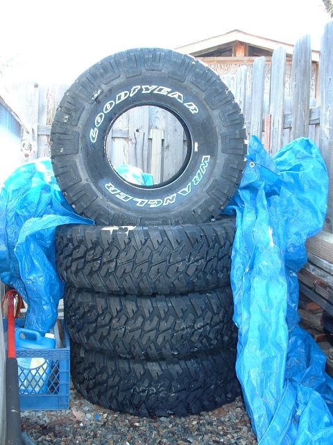1991 Ford Bronco - Four unmounted Goodyear Wrangler mud-terrain tires, three stacked flat with one stood on edge showing the raised white lettering and aggres…