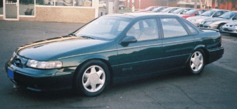 1998 Ford Taurus - A dark green sedan is parked on a street, with buildings in the background.