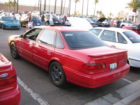 1998 Ford Taurus - A red sedan with a spoiler is parked at an outdoor event with other vehicles and people.