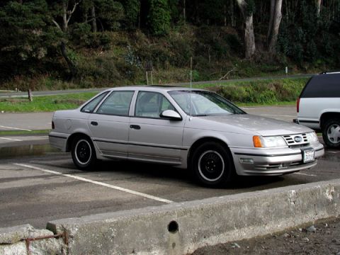 1998 Ford Taurus - A silver sedan is parked in a lot, with a white SUV visible to its right.