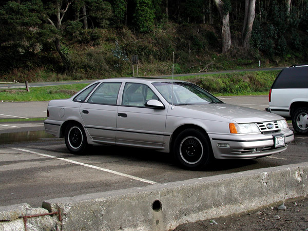 Silver sedan parked in a lot, next to a white SUV. It has black wheels and a sunroof