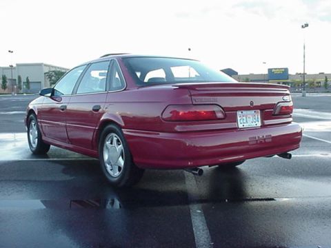 1998 Ford Taurus - A maroon sedan with a rear spoiler is parked on a wet asphalt surface, showing its rear 3/4 view.