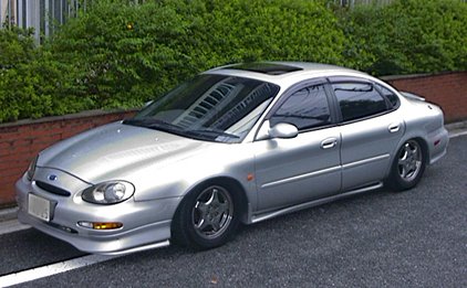 1998 Ford Taurus - A silver 1998 Ford Taurus is parked on the side of the road with a hedge and brick wall behind it.