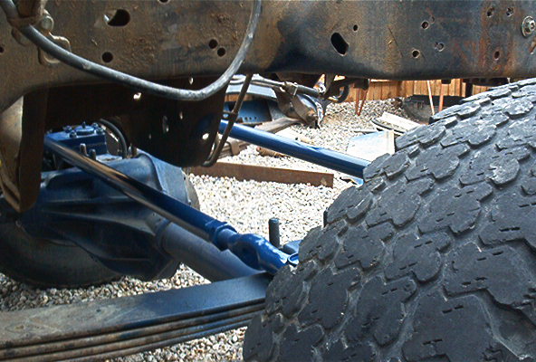 Close-up of a vehicle's rusty frame, leaf springs, and rear axle with blue suspension components. Large tire in foreground