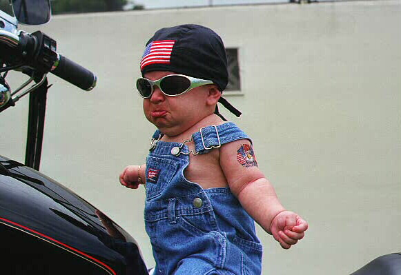 Baby in overalls and sunglasses sits on a motorcycle, wearing a bandana with an American flag patch