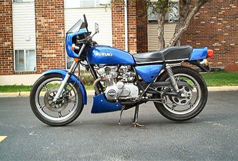 1985 Ford Crown Victoria LTD - A blue Suzuki motorcycle is parked on asphalt in front of a brick building with a tree nearby.