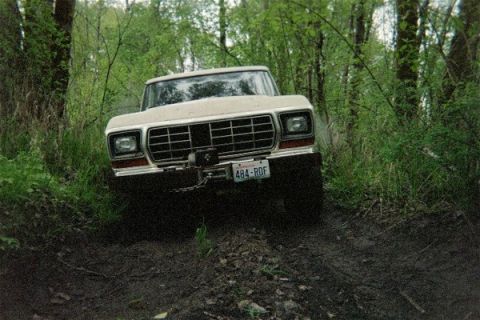 1978 Ford Bronco - A 1978 Ford Bronco navigates a muddy off-road path surrounded by trees.