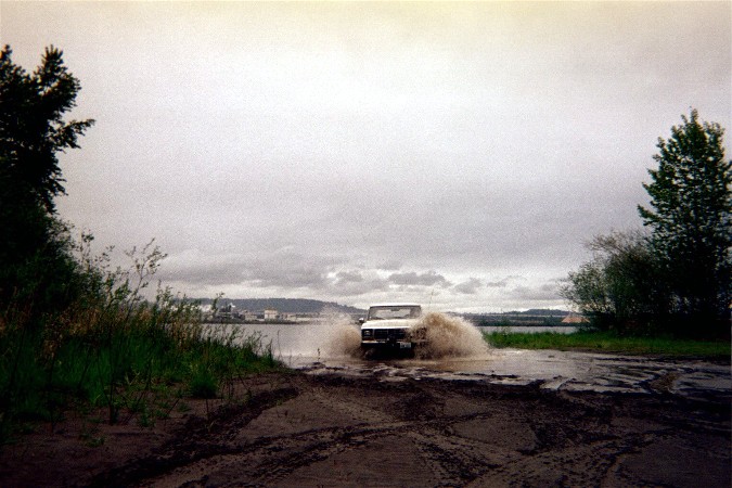 1978 Ford Bronco driving through a shallow body of water, kicking up a large splash