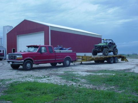 1992 Ford Bronco - A red Ford pickup truck tows a trailer carrying a green off-road vehicle in front of a large red barn.