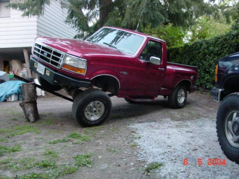 1992 Ford F-150 - A red 1992 Ford F-150 pickup truck is propped up on blocks, with another dark-colored vehicle visible to the right.