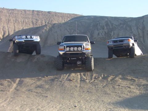 2000 Ford F-450 - Three trucks are parked on sand dunes under a clear sky. The middle truck is a dark color with off-road lights.