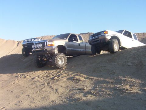 2000 Ford F-450 - Three lifted trucks, including a 2000 Ford F-450, navigate sandy terrain under a clear blue sky.