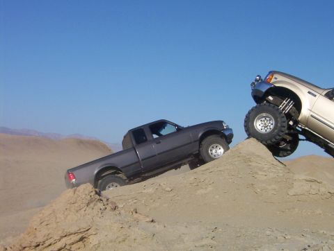 2000 Ford F-450 - Two pickup trucks navigate challenging terrain on a sunny day, with one truck climbing a steep sand dune.