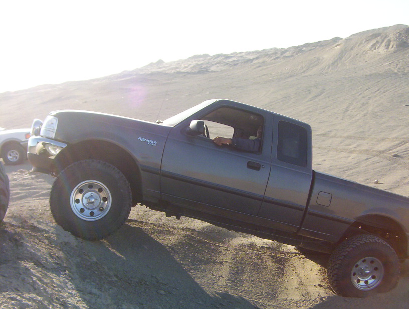 Dark gray Ford Ranger pickup truck with lifted suspension driving on a sand dune. Another white vehicle is partially visible to the left