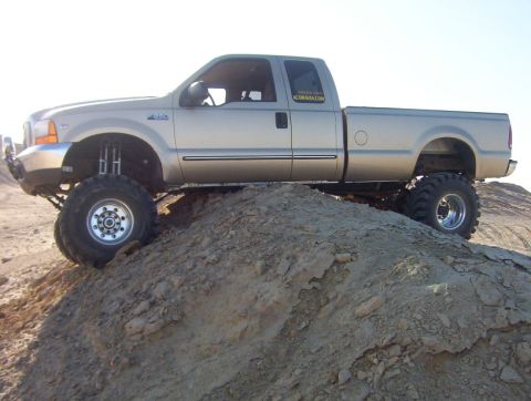 2000 Ford F-450 - A lifted silver Ford F-450 pickup truck is positioned on a dirt incline under a clear sky.