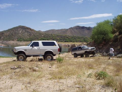 1993 Ford Bronco - Two Ford Broncos are seen off-roading near a body of water with mountains in the background.