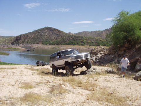 1993 Ford Bronco - A two-tone SUV navigates rocky terrain near a lake, with another vehicle visible in the background.