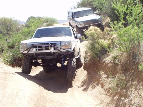 1993 Ford Bronco - Two SUVs navigate a steep, dusty off-road trail with sparse desert vegetation.