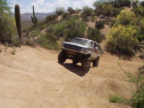 1993 Ford Bronco - A lifted silver SUV navigates a sandy, off-road trail in a desert environment, with cacti visible.