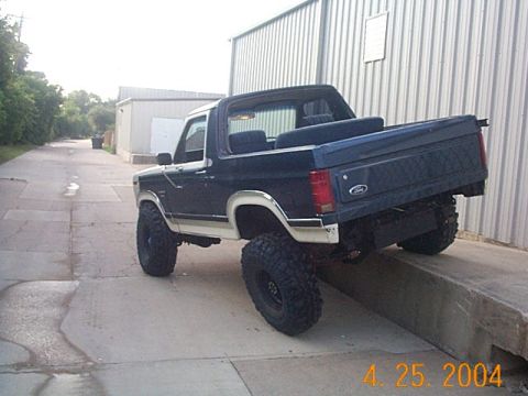 1986 Ford Bronco - Lifted Ford Bronco with aftermarket wheels and tires sits on a concrete incline next to a metal building.
