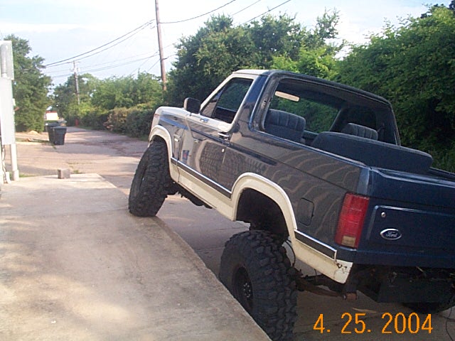 Lifted blue and white topless Ford Bronco II flexing a front tire up on a curb