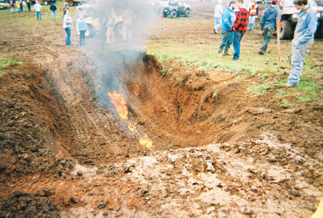 Flames and smoke in a churned mud pit with spectators and trucks around it
