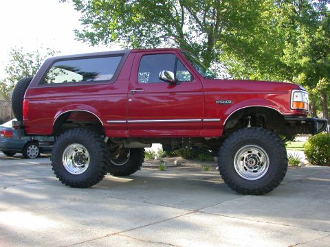 1996 Ford Bronco - A clean red 1990s full-size Ford Bronco photographed in profile on a concrete driveway, showing its hardtop, 'BRONCO' fender lettering and…