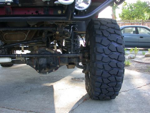 1996 Ford Bronco - Close-up of an off-road vehicle's front suspension and tire, with a sedan visible in the background.