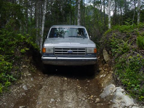 1987 Ford Bronco - A silver Ford Bronco navigates a rough, wooded trail.