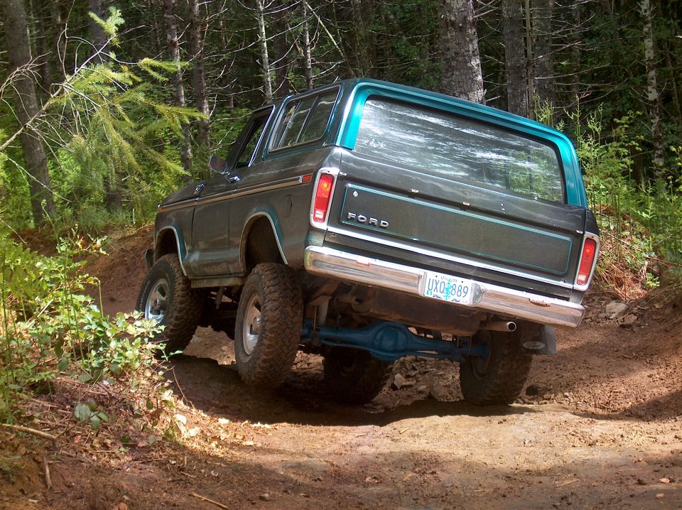 Lifted teal Ford Bronco from the rear, climbing a dirt off-road trail through forest