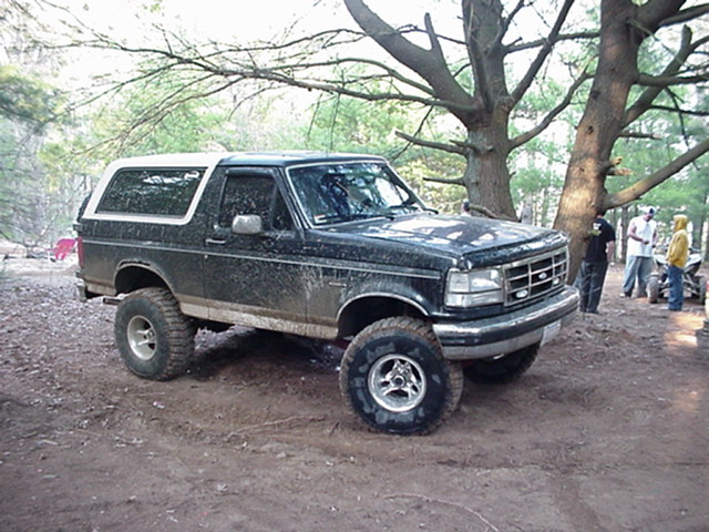 Dark green Ford Bronco with a white top, lifted suspension, and mud splatters, parked on a dirt trail