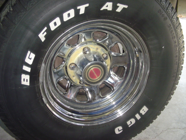 Close-up of a chrome wheel with a 'BIG FOOT AT' tire. Center cap has a red and silver emblem