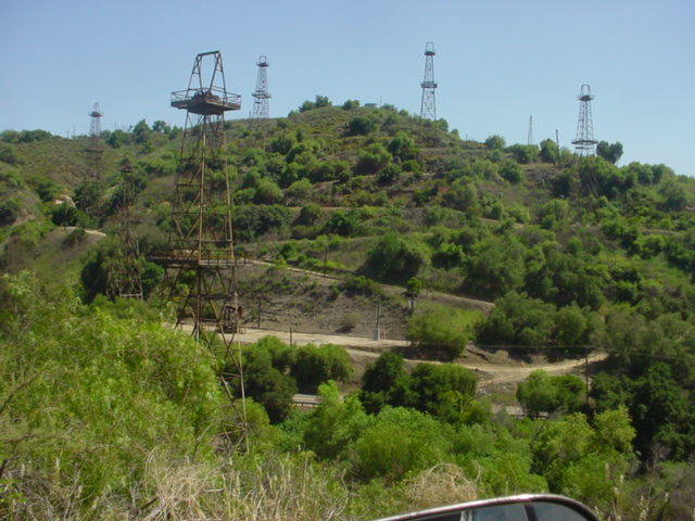 Oil derricks stand on a green, hilly landscape under a clear blue sky