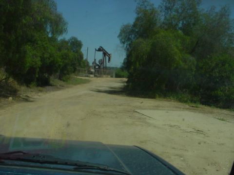 1988 Ford Bronco - A dirt road winds through a wooded area towards an active oil pumpjack under a clear blue sky.