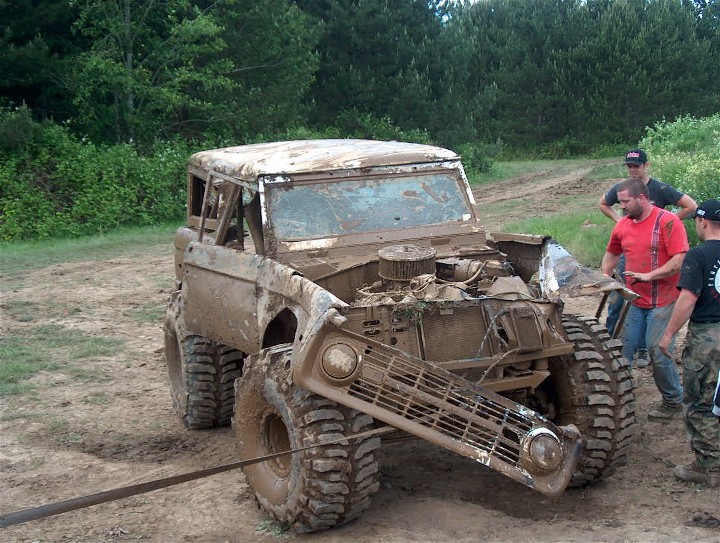 Muddy 1979 Ford Bronco with grille detached, hood open, and engine exposed. Three men stand nearby