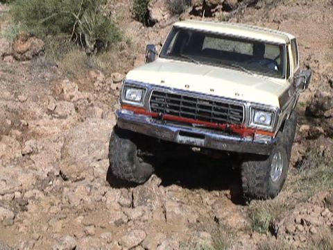 1979 Ford Bronco - Elevated front view of a cream-colored Ford Bronco with FORD hood lettering picking its way down a rocky desert descent, front tires stradd…