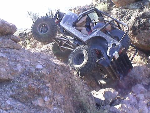 1979 Ford Bronco - Overhead view of a bare-metal tube-frame rock buggy climbing a steep rocky ledge, front left tire lifted high off the rock while the driver…