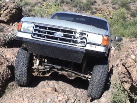 1979 Ford Bronco - Low front view of a lifted two-tone silver and blue Ford truck flexing on rocky desert trail, right front tire drooped into a hole and left…