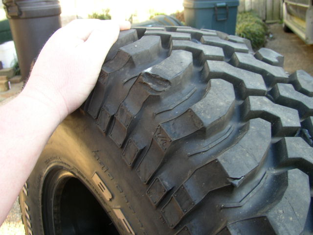 Close-up of a large, knobby off-road tire with a hand resting on it. Some tread damage is visible