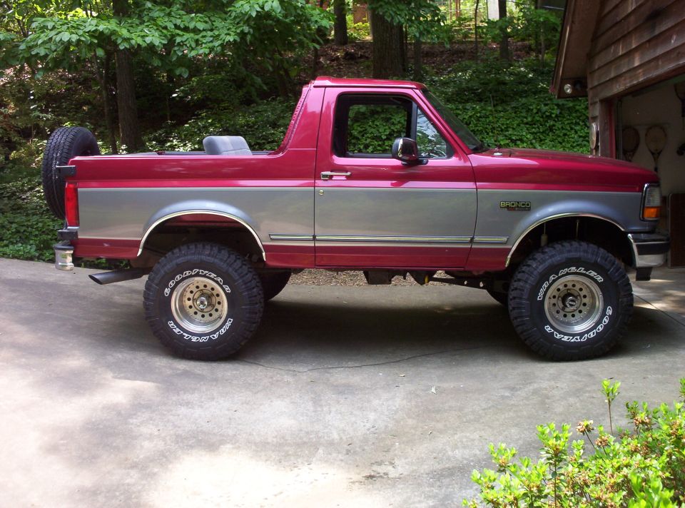 Red and silver Ford Bronco parked outside a garage. Has a spare tire on the back