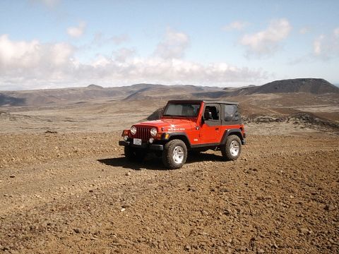1996 Ford Bronco - A red Jeep Wrangler Rubicon is parked on a dirt road with a vast, rocky, and mountainous terrain in the background under a partly cloudy sk…