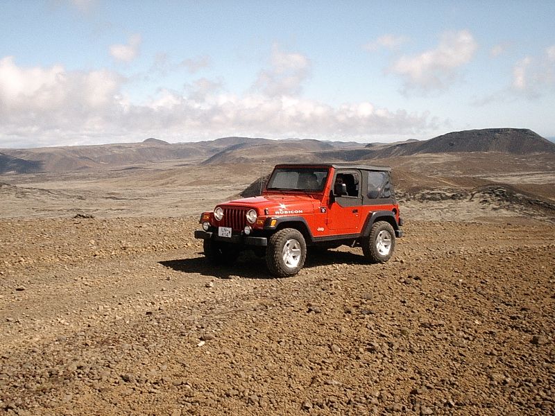 Red Jeep Wrangler Rubicon soft top on gravel terrain with hills behind