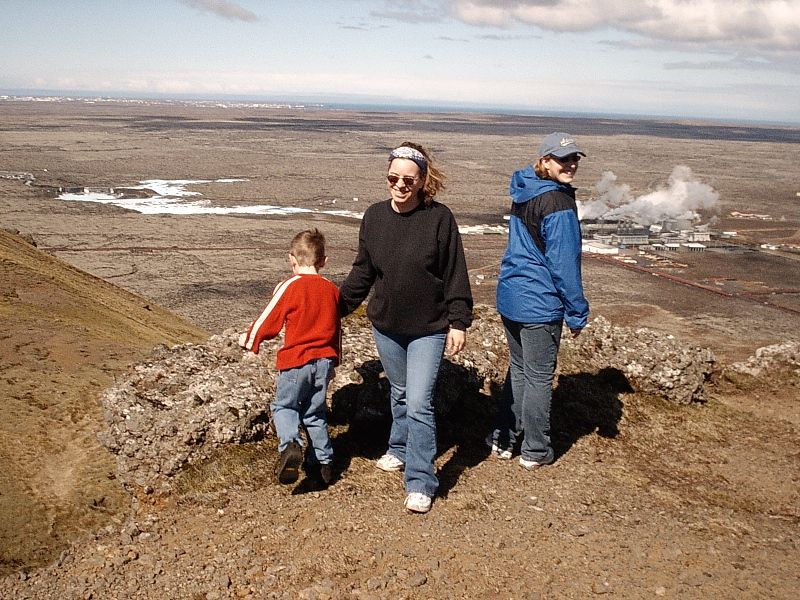 Woman, boy, and person on rocky hill overlooking plain with distant steam plumes