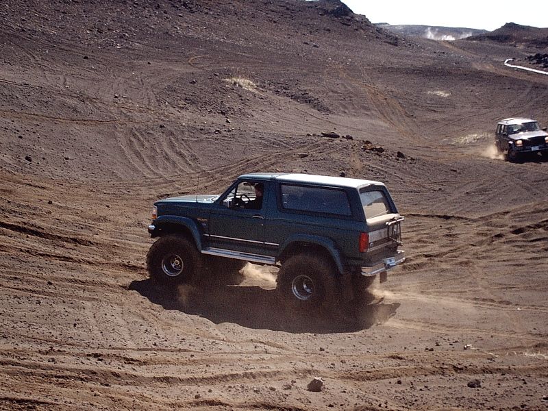 Green two-door Bronco with white top driving on gray volcanic dirt, kicking up dust