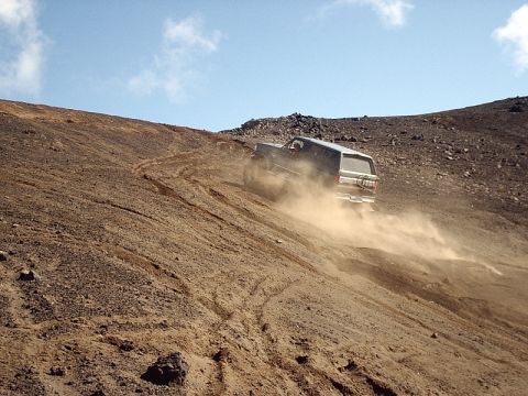 1996 Ford Bronco - A dark SUV navigates a dusty, off-road incline under a clear blue sky.