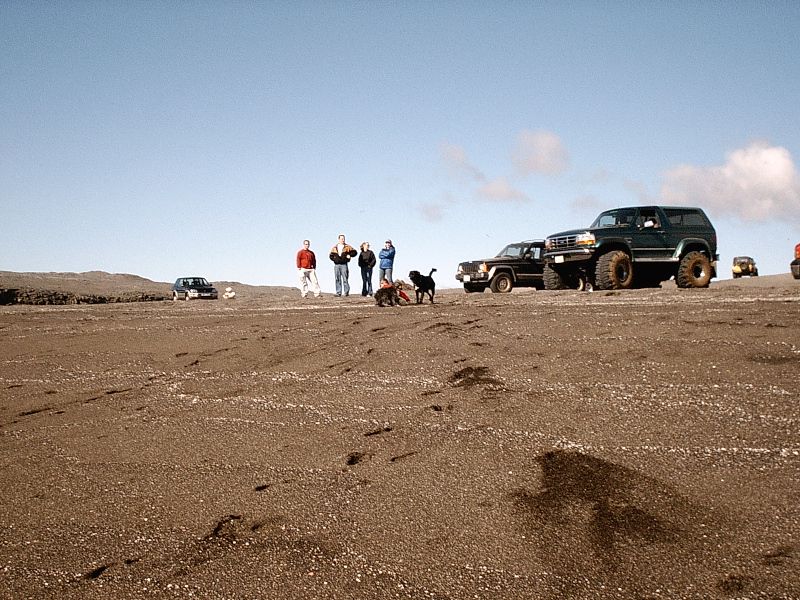 Dark green lifted Ford Bronco with big tires on gray sand plain, people and dog nearby