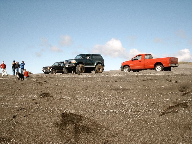 Lifted dark green Bronco on sand between a wagon and red pickup, people and dog at left.